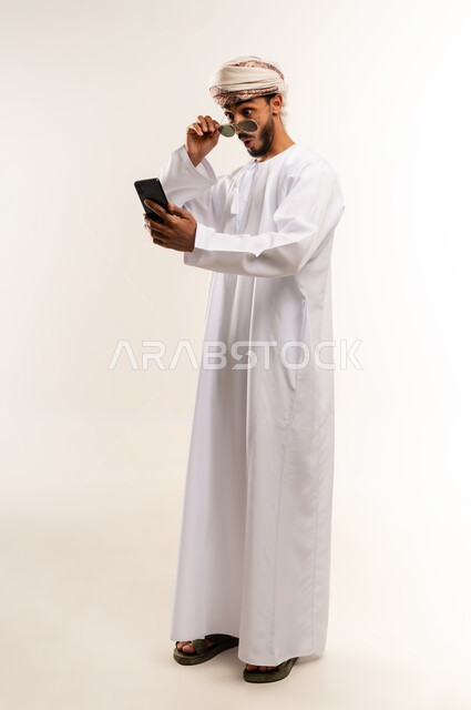 استخدام جهاز تقني حديث ومتطور, توثيق اللحظات السعيدة عبر الهاتف المحمول, بورتريه لشاب عربي خليجي عماني يرتدي الدشداشة والعمامة يضع نظارة شمسية ويقوم بالتقاط صورة السيلفي التذكارية عبر الجوال, الجسم بالطول الكامل, خلفية بيضاء .