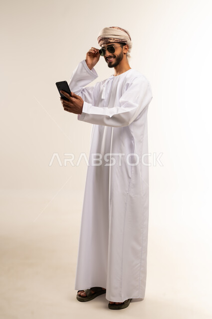 توثيق اللحظات السعيدة عبر الهاتف المحمول, استخدام جهاز تقني حديث ومتطور, بورتريه لشاب عربي خليجي عماني يرتدي الدشداشة والعمامة يضع نظارة شمسية ويقوم بالتقاط صورة السيلفي التذكارية عبر الجوال, الجسم بالطول الكامل, خلفية بيضاء .
