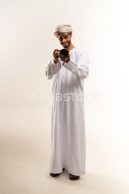 توثيق اللحظات السعيدة, الشغف بممارسة الهواية المفضلة، بورتريه لشاب عربي خليجي عماني مبتسم يرتدي الدشداشة والكمه يقوم بالتقاط صور تذكارية عبر كاميرا فوتوغرافية، تعابير الفرح والسرور، استخدام كاميرات رقمية عالية الدقة، صورة الجسم بالطول الكامل، خلفية بيضاء .