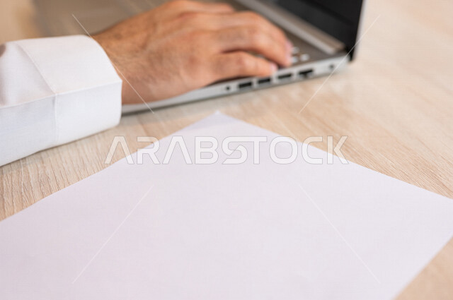 A Saudi Arabian Gulf man sitting on a laptop in front of him is a set of blank papers, conducting business through the laptop, entering data through the keyboard, text contents