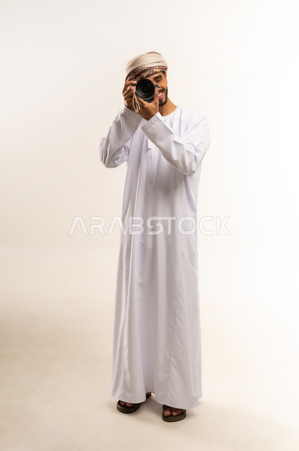 إتقان واحتراف مهنة التصوير، بورتريه لشاب عربي خليجي عماني مبتسم يرتدي الدشداشة والعمامة يقوم بالتقاط صور تذكارية عبر كاميرا فوتوغرافية، تعابير الفرح والسرور، استخدام كاميرات رقمية عالية الدقة، صورة الجسم بالطول الكامل، خلفية بيضاء .