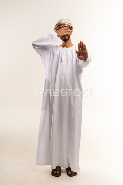 طلب التوقف وعدم الاقتراب, مفهوم الرفض والامتناع والانزعاج, بورتريه لرجل عربي خليجي عماني يرتدي الدشداشة والعمامة يضع يده على عيونه يمد اليد الأخرى نحو الأمام مع رفع الكف بإيماءات النهي عن فعل امر ما, تعابير الحزم والجدية, صورة الجسم بالطول الكامل، خلفية بيضاء .