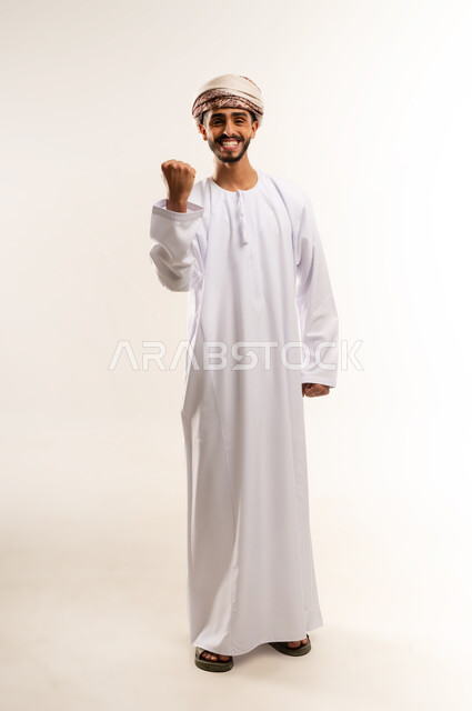 تعابير السرور والسعادة، مفهوم الانجاز وتحقيق الاهداف، القوة والتحدي والحماس، بورتريه لرجل عربي خليجي عماني يرتدي الدشداشة والعمامة يرفع قبضتيه نحو الأعلى بإيماءات التشجيع، صورة الجسم بالطول الكامل، خلفية بيضاء .