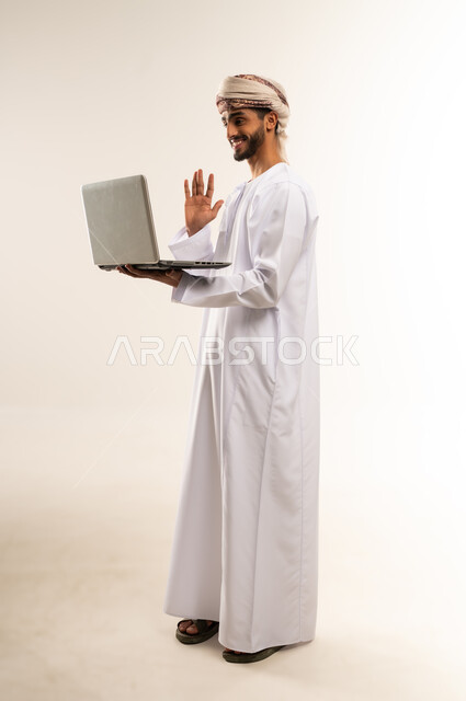 Live broadcasting, using modern technologies and advanced devices, the concept of facilitating work and the ability to complete tasks remotely. Portrait of a young Gulf Arab Omani man wearing a dishdasha and turban, holding a laptop, showing signs of joy and happiness. Full-length body, white background.
