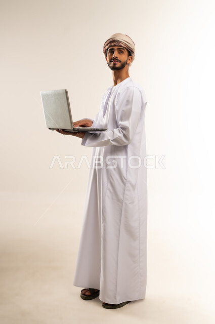 Using modern technologies and advanced devices, the concept of facilitating work and the ability to complete tasks remotely. Portrait of a young Gulf Arab Omani man wearing a dishdasha and turban, holding a laptop, showing signs of joy and happiness. Full-length body, white background.