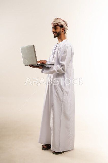 مفهوم تسهيل الاعمال والقدرة على إنجاز المهام عن بعد، استخدام التقنيات الحديثة والأجهزة المتطورة ، بورتريه لشاب عربي خليجي عماني يرتدي الدشداشة والعمامة يمسك بيده حاسوب محمول تظهر عليه علامات الفرح والسرور، الجسم بالطول الكامل، خلفية بيضاء