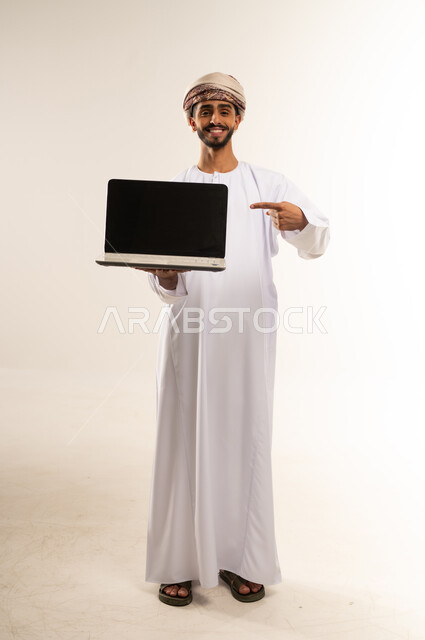 Using advanced technology for digital marketing, displaying computer applications and services. Portrait of an Omani Gulf Arab man wearing a dishdasha and turban, holding a laptop with a blank black screen. Full-length body, white background.