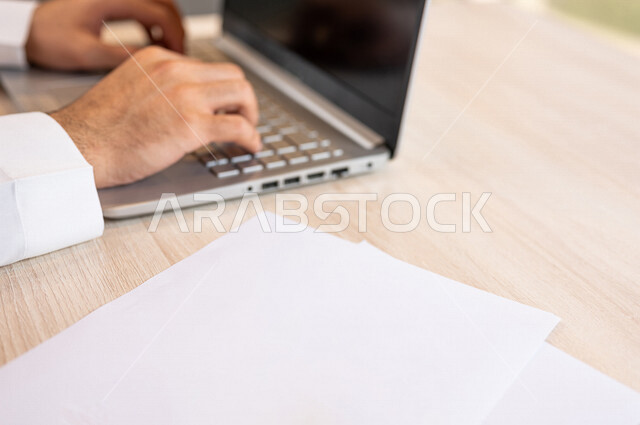 رجل عربي خليجي سعودي يجلس على الحاسوب المحمول وامامه مجموعة من الأوراق الفارغة، ادارة الاعمال من خلال الحاسوب المحمول، ادخال البيانات من خلال لوحة المفاتيح،  محتويات نصية