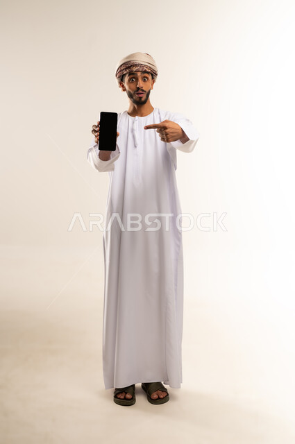 Using advanced technology, displaying mobile applications and services, the concept of digital and electronic marketing. Portrait of an Omani Gulf Arab man wearing a dishdasha and turban, holding a mobile phone with a blank black screen and pointing at it with his index finger. Full-length body image, white background.