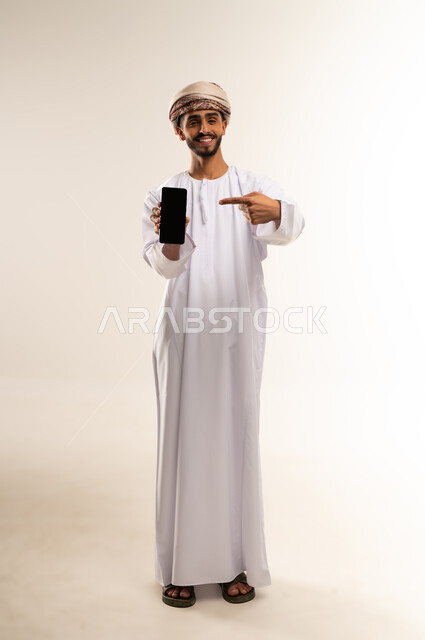 Mobile application and service presentation, digital and e-marketing concept, portrait of an Omani Gulf Arab man wearing a dishdasha and turban, holding a mobile phone with a blank black screen and pointing at it with his index finger. Use of advanced technology, full-length body image, white background.