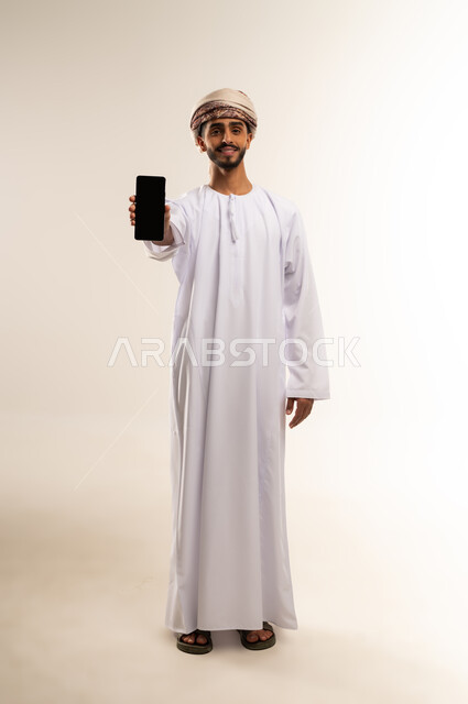 مفهوم التسويق الرقمي و الالكتروني، عرض تطبيقات وخدمات الهاتف المحمول، بورتريه لرجل عربي خليجي عماني يرتدي الدشداشة والعمامة يحمل بيده جوال بشاشة سوداء فارغة، استخدام التكنولوجيا المتطورة، صورة الجسم بالطول الكامل، خلفية بيضاء.