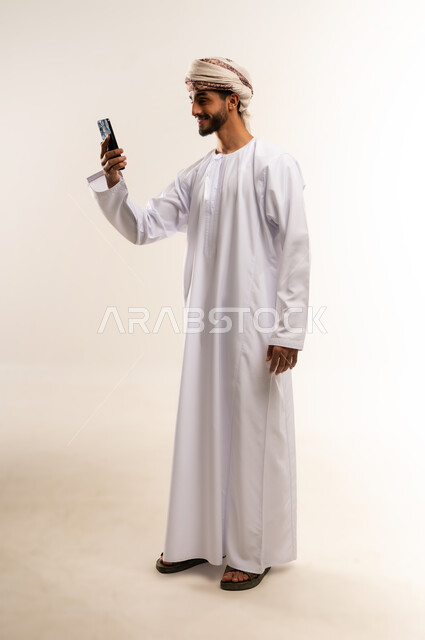 The concept of facilitating business and the ability to complete tasks remotely, using modern technologies and advanced devices, a portrait of a young Arab Gulf Omani man wearing a dishdasha and turban, holding a mobile phone, showing signs of joy and happiness, full body, white background.