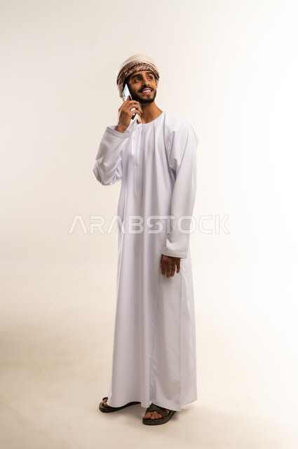 تبادل الأحاديث مع الأهل والأصدقاء، إجراء مكالمة هاتفية باستخدام الهاتف المحمول، بورتريه بالطول الكامل لرجل عربي خليجي عماني يرتدي الدشداشة والعمامة يقوم بالتحدث بهاتفه المحمول بتعابير الفرح والسرور، خلفية بيضاء .