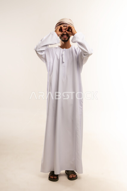 حب الاستكشاف و المغامرة, رؤية الاشياء البعيدة و تقريبها, بورتريه لشاب عربي خليجي عماني يرتدي الدشداشة والعمامة مبتسم يحمل بيده منظار ثنائي العينين و يينظر الى شيئ ما, ايماءات الاستمتاع و السرور, صورة الجسم بالطول الكامل, خلفية بيضاء .