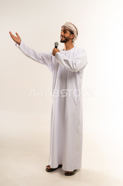 الاستمتاع بممارسة المواهب الفنية، تسجيل مقطع صوتي غنائي, قضاء وقت ممتع بأداء العروض الصوتية، بورتريه لشاب عربي خليجي عماني يرتدي الدشداشة والعمامة  يمسك بيده المايكروفون ينظر الى شي ما بايماءات السعادة والسرور, الجسم بالطول الكامل, خلفية بيضاء .