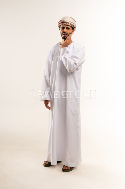 طلب العلاج والمساعدة، تسوس الاضراس و التهاب اللثه، بورتريه لرجل عربي خليجي عماني يرتدي الدشداشة والعمامة يضع يده على خده ويغمض عينيه بايماءات الشعور بألم الأسنان، الحاجة لزيارة الطبيب، الجسم بالطول الكامل، خلفية بيضاء .