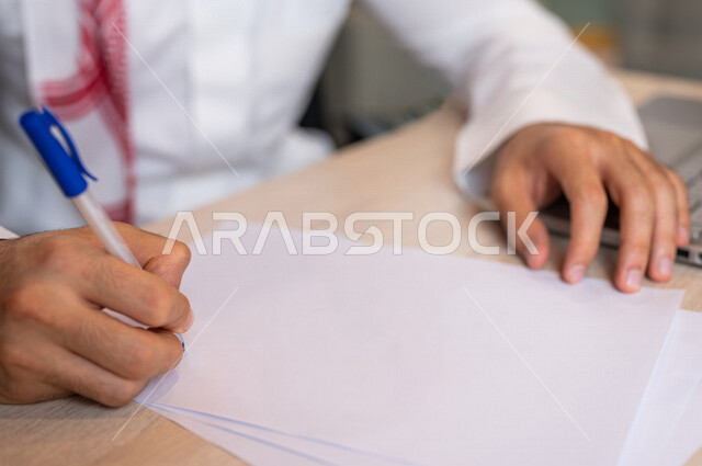A Saudi Arabian Gulf man sitting in front of a laptop, blank white papers, a secretary managing paperwork, office work