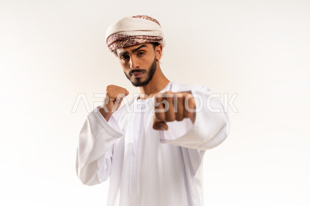 ممارسة رياضة الدفاع عن النفس، إتقان فن الملاكمة، بورتريه مقرب لرجل عربي خليجي عماني يرتدي الدشداشة التقليدية والعمامة يرفع قبضات يديه ينظر إلى الكاميرا بإيماءات القوة، زيادة قدرة العضلات على التحمل، تعزيز صحة العظام والمفاصل، الجسم بالطول الكامل، خلفية بيضاء .