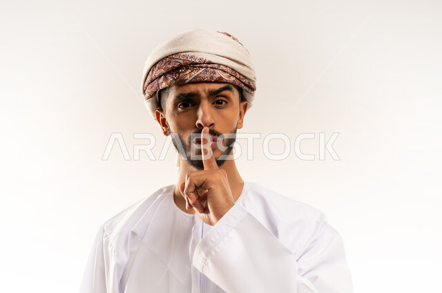 Request to stop talking and be silent, prohibition, admonition and listening, index finger in front of mouth with serious gestures, order for complete silence, close-up portrait of an Arab Gulf Omani man wearing a dishdasha and turban looking at the camera with angry gestures, full body, white background.
