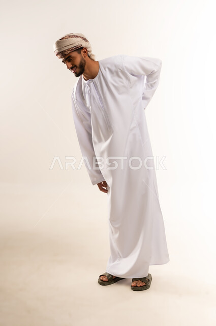 الأوجاع والآلام العصبية، طلب المساعده والحاجه للعلاج، التهاب العضلات والمفاصل، بورتريه لشاب عربي خليجي عماني يرتدي الدشداشة والعمامة يضع يده على ظهره بايماءات الألم والتعب والأرهاق، حالة اسعافية طارئة، صورة الجسم الطول الكامل, خلفية بيضاء .