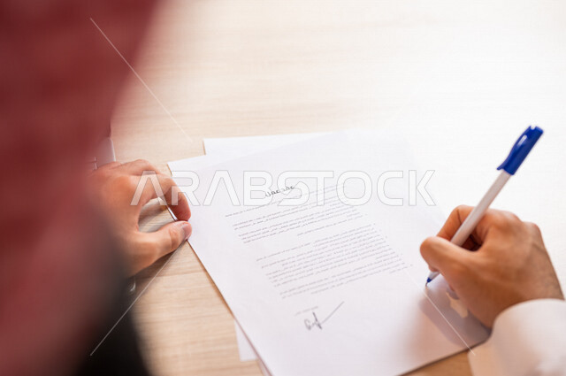 صورة مقربة ليد رجل عربي خليجي سعودي يقوم بقراءة البنود الخاصة بالعمل، التحقق والتدقيق في شروط عقد العمل، الموافقة على الشروط، التوقيع على العقود