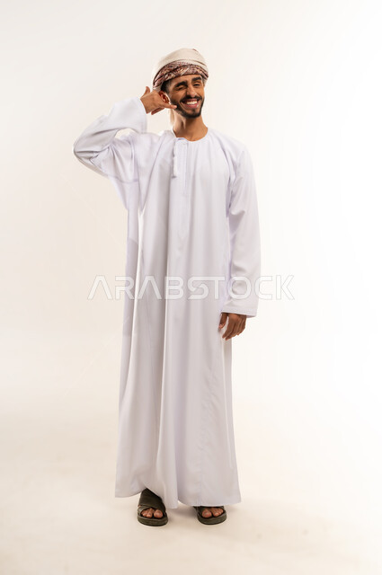 مفهوم تبادل المعلومات، حركة يد تدل على الاتصال، الاهتمام بالمظهر الخارجي والوسامة، بورتريه لرجل عربي خليجي عماني يرتدي الدشداشة والعمامة يرفع يده بايماءات الرد على المكالمات الهاتفية، تلقي أخبار سارة عبر الجوال، الجسم بالطول الكامل، خلفية بيضاء.