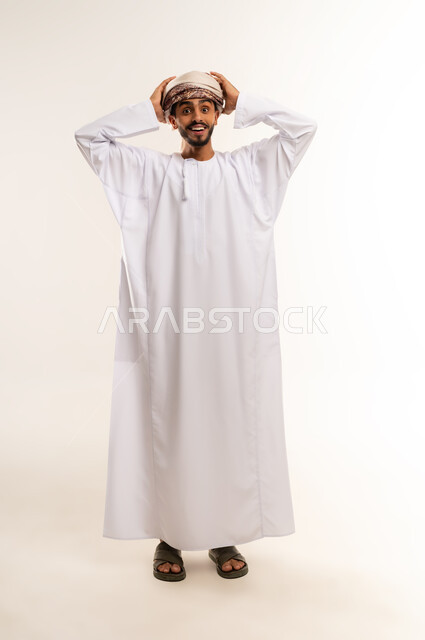 مفهوم الاستغراب والانبهار ، الشعور بالدهشة والصدمة والذهول, التفاجؤ من شيء ما، بورتريه لشاب عربي خليجي عماني مبتسم يرتدي الدشداشة والعمامة يضع يديه على رأسه بإيماءات التعجب, صورة الجسم بالطول الكامل, خلفية بيضاء