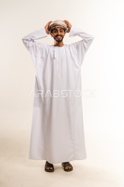 التفاجؤ من شيء ما، الشعور بالدهشة والصدمة والذهول, بورتريه لشاب عربي خليجي عماني مبتسم يرتدي الدشداشة والعمامة  يضع يديه على رأسه بإيماءات التعجب, صورة الجسم بالطول الكامل, خلفية بيضاء