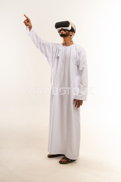 الاشارة الى شئ ما، التطور التقني والتكنولوجي, الاندماج ومحاكاة العالم الخيالي والنظر الى الافلام الواقعيه، المتعة و التسلية بالمشاهدة عبر نظارة الواقع الافتراضي، بورتريه لشاب عربي خليجي عماني يرتدي الدشداشه والكمه ينظر من خلال نظارات ثلاثية الأبعاد (الفي ار), خلفيه بيضاء .
