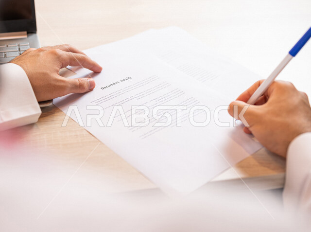 رجل عربي خليجي سعودي يجلس على الحاسوب المحمول وأمامه وثيقة، انجاز مهام العمل من خلال الحاسوب المحمول، سكرتير يدير الاعمال الورقية، التوقيع على الوثيقة