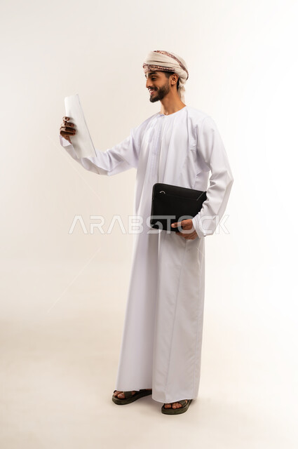 Caring about elegance and outward appearance, going to work energetically, feeling happy to achieve the company's goals, portrait of a smiling Omani Gulf Arab man wearing a dishdasha and a kummah, looking to the side with happy gestures, carrying a black handbag and work papers, full body image, white background.