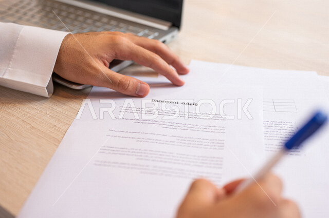 رجل عربي خليجي سعودي يجلس على الحاسوب المحمول وأمامه وثيقة، انجاز مهام العمل من خلال الحاسوب المحمول، سكرتير يدير الاعمال الورقية، التوقيع على الوثيقة