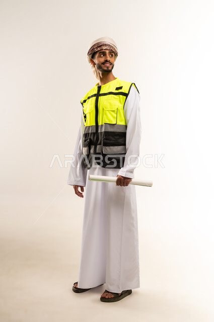 النظر جانباً بإيماءات السعادة والسرور، العمل في مجال الهندسة، الإشراف على مخططات المشاريع الهندسية، مفهوم الهندسة والانشاءات المعمارية، بورتريه لمهندس عربي خليجي عماني يرتدي الدشداشة والكمة وسترة الحماية يمسك بيديه المخطط، مهن ووظائف للشباب في سلطنة عمان، صورة الجسم بالطول الكامل ،خلفية بيضاء .