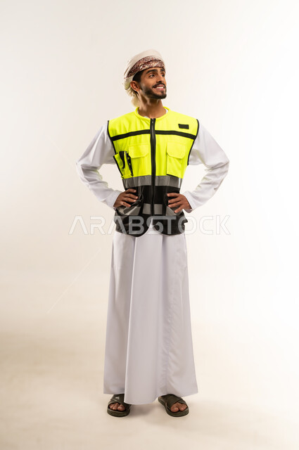 مفهوم الهندسة والانشاءات المعمارية, العمل في القطاع الهندسي, الإعمار بسواعد أبناء الوطن, النظر إلى الكاميرا ما بإيماءات السعادة والسرور، بورتريه لمهندس عربي خليجي عماني يرتدي الدشداشة والكمة مع سترة الحماية يقف باستقامة واعتدال, الجسم بالطول الكامل, خلفية بيضاء .