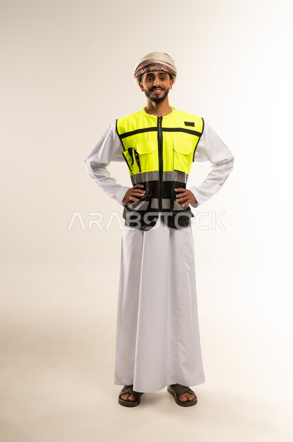 المتابعة والإشراف على المشاريع الهندسية,  العمل في القطاع الهندسي, الإعمار بسواعد أبناء الوطن, النظر إلى الكاميرا ما بإيماءات السعادة والسرور، بورتريه لمهندس عربي خليجي عماني يرتدي الدشداشة والكمة مع سترة الحماية يقف باستقامة واعتدال, الجسم بالطول الكامل, خلفية بيضاء .