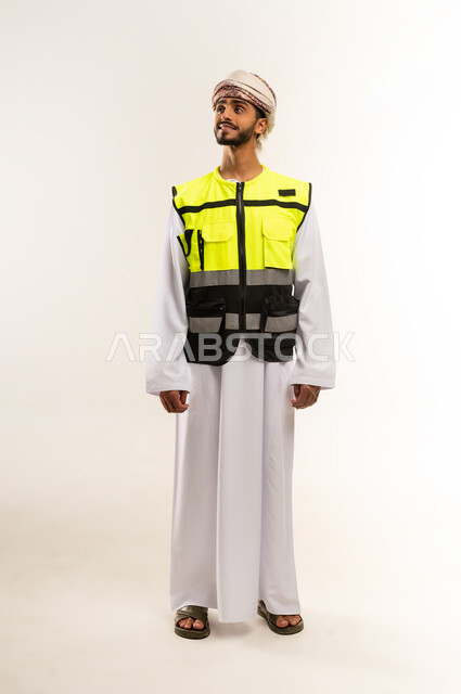 العمل في القطاع الهندسي, الإعمار بسواعد أبناء الوطن, النظر إلى شئ ما بإيماءات السعادة والسرور، المتابعة والإشراف على المشاريع الهندسية, بورتريه لمهندس عربي خليجي عماني يرتدي الدشداشة والكمة مع سترة الحماية يقف باستقامة واعتدال, الجسم بالطول الكامل, خلفية بيضاء .