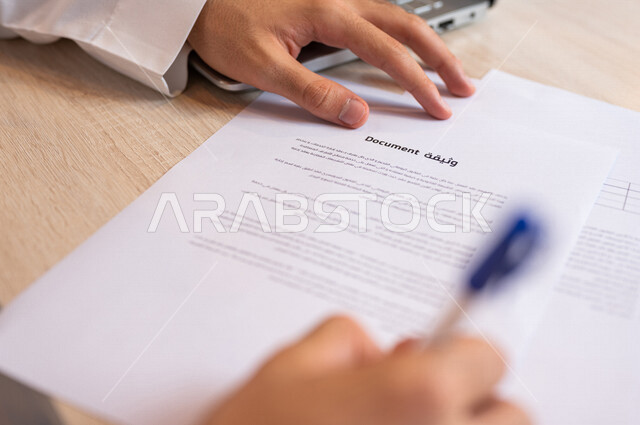 صورة مقربة ليد رجل عربي خليجي سعودي يقوم بقراءة البنود الخاصة بالعمل، التحقق والتدقيق في شروط عقد العمل، الموافقة على الشروط، التوقيع على العقود