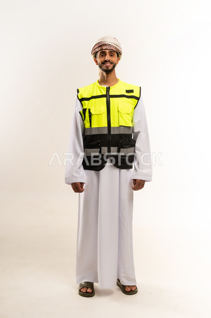 Looking at the camera with gestures of happiness and pleasure, working in the engineering sector, reconstruction with the hands of the sons of the nation, following up and supervising engineering projects, portrait of an Arab Gulf Omani engineer wearing a dishdasha and a kummah with a protective jacket, standing straight and balanced, full body, white background.