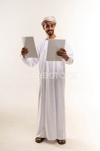 Daily task recording tools, using white paper to record notes and important information, portrait of an Arab Gulf Omani man wearing a dishdasha and a kuma holding work papers with gestures of concentration and contemplation, full-length body, white background