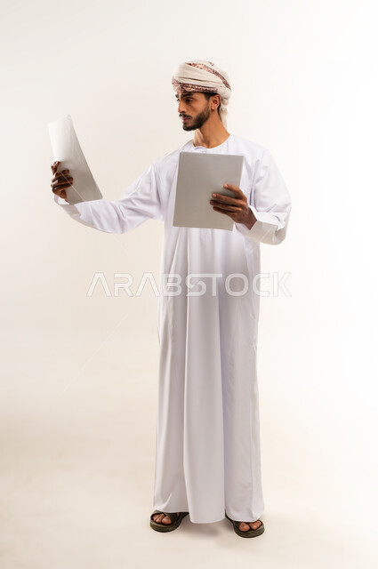 استخدام الورق الأبيض في تسجيل الملاحظات والمعلومات المهمة, بورتريه لرجل عربي خليجي عماني يرتدي الدشداشة والكمة يمسك بيده اوراق العمل بإيماءات التركيز والتمعن، أدوات تدوين المهام اليومية، الجسم بالطول الكامل، خلفية بيضاء
