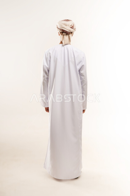 ارتداء الملابس العمانية التقليدية ، الاهتمام بالأناقة والمظهر الخارجي، الوقوف باستقامة واعتدال، بورتريه من الخلف لشاب عربي خليجي عماني مبتسم يرتدي الدشداشة و الكمه ينظر الى الكاميرا بإيماءات السرور والسعادة، صورة الجسم بالطول الكامل, خلفية بيضاء