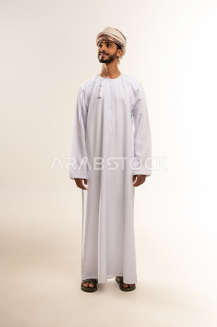 الاهتمام بالأناقة والمظهر الخارجي، مفهوم القوة والرجولة، بورتريه  لرجل عربي خليجي عماني يرتدي الدشداشه والعمامة يقوم بالنظر الى مكان ما جانبا، ايماءات الثقة بالنفس، صورة الجسم بالطول الكامل ، خلفية بيضاء .