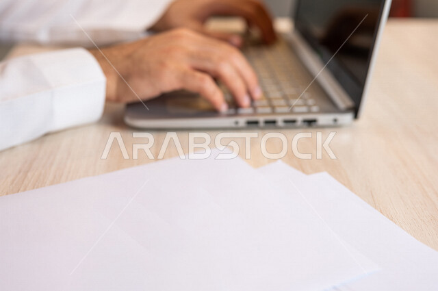 رجل عربي خليجي سعودي يجلس على الحاسوب المحمول وامامه مجموعة من الأوراق الفارغة، ادارة الاعمال من خلال الحاسوب المحمول، ادخال البيانات من خلال لوحة المفاتيح،  محتويات نصية