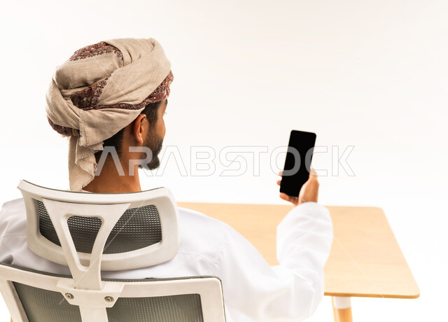 اجراء مكالمات الفيديو عبر الجوال ، الاستفادة من التقنيات الحديثة في القطاع المكتبي والإداري، مفهوم إدارة العمل عن بعد، بورتريه من الخلف لرجل عربي خليجي عماني يرتدي الدشداشه والعمامه يجلس على طاولة المكتب ينظر إلى مكان ما بإيماءات الاندماج، خلفية بيضاء .