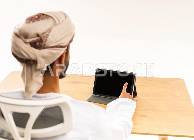 The concept of remote work management, supervising workflow via tablet, utilizing modern technologies in the office and administrative sector, a portrait from the back of an Arab Gulf Omani man wearing a dishdasha and turban sitting at an office table looking somewhere with gestures of integration, white background.