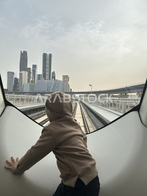 التطوير والتقدم الحضاري وتحسين البنية التحتيه، شبكة السكك الحديدية للنقل السريع، محطة مترو الرياض، وسائل النقل والمواصلات الحديثة في المملكة العربية السعودية، معلم سياحي مشهور، صورة من الخلف لولد عربي خليجي سعودي يشاهد معالم العاصمة من داخل القطار