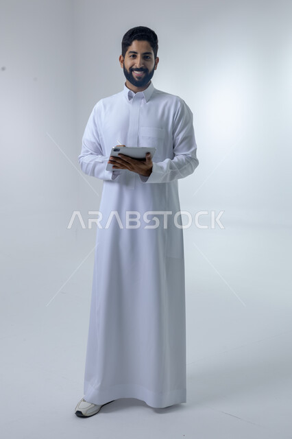 Using modern advanced technology in practical life via tablet, expressions and gestures of integration, portrait of a smiling Saudi Arabian Gulf young man wearing traditional dress working on a tablet, full body image, white background