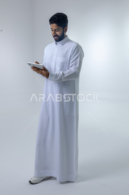 استخدام التقنية الحديثه المتطورة في الحياة العملية عبر الجهاز اللوحي، تعابير وايماءات الاندماج، بورتريه لشاب عربي خليجي سعودي مبتسم يرتدي الثوب التقليدي يقوم بالعمل على جهازالتابلت، صورة الجسم بالطول الكامل، خلفية بيضاء