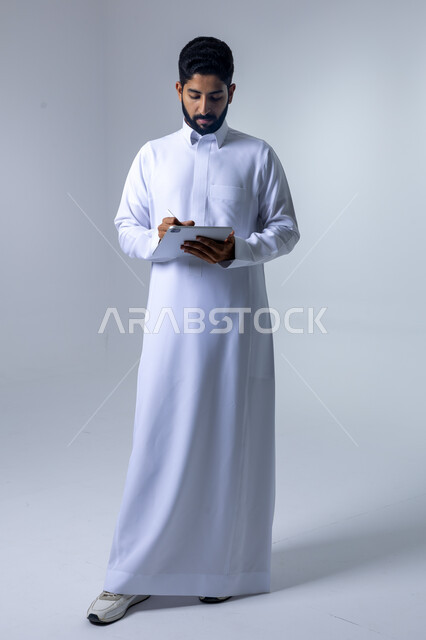 استخدام التقنية الحديثه المتطورة في الحياة العملية عبر الجهاز اللوحي، تعابير وايماءات الاندماج، بورتريه لشاب عربي خليجي سعودي مبتسم يرتدي الثوب التقليدي يقوم بالعمل على جهازالتابلت، صورة الجسم بالطول الكامل، خلفية بيضاء