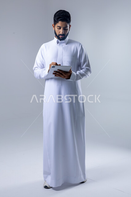 Using modern advanced technology in practical life via tablet, expressions and gestures of integration, portrait of a smiling Saudi Arabian Gulf young man wearing traditional dress working on a tablet, full body image, white background